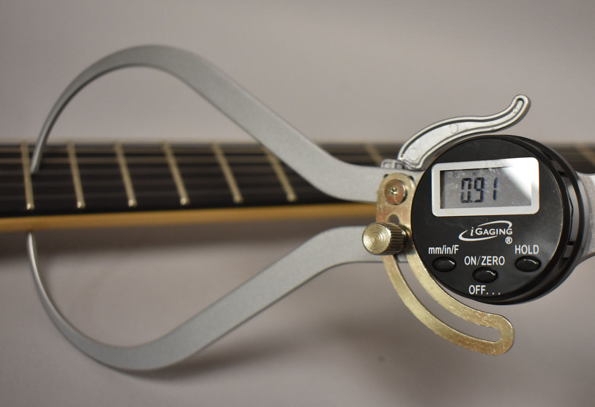 A digital caliper measures the fret height on a dark fretboard with light frets, displaying "0.91" on its screen.