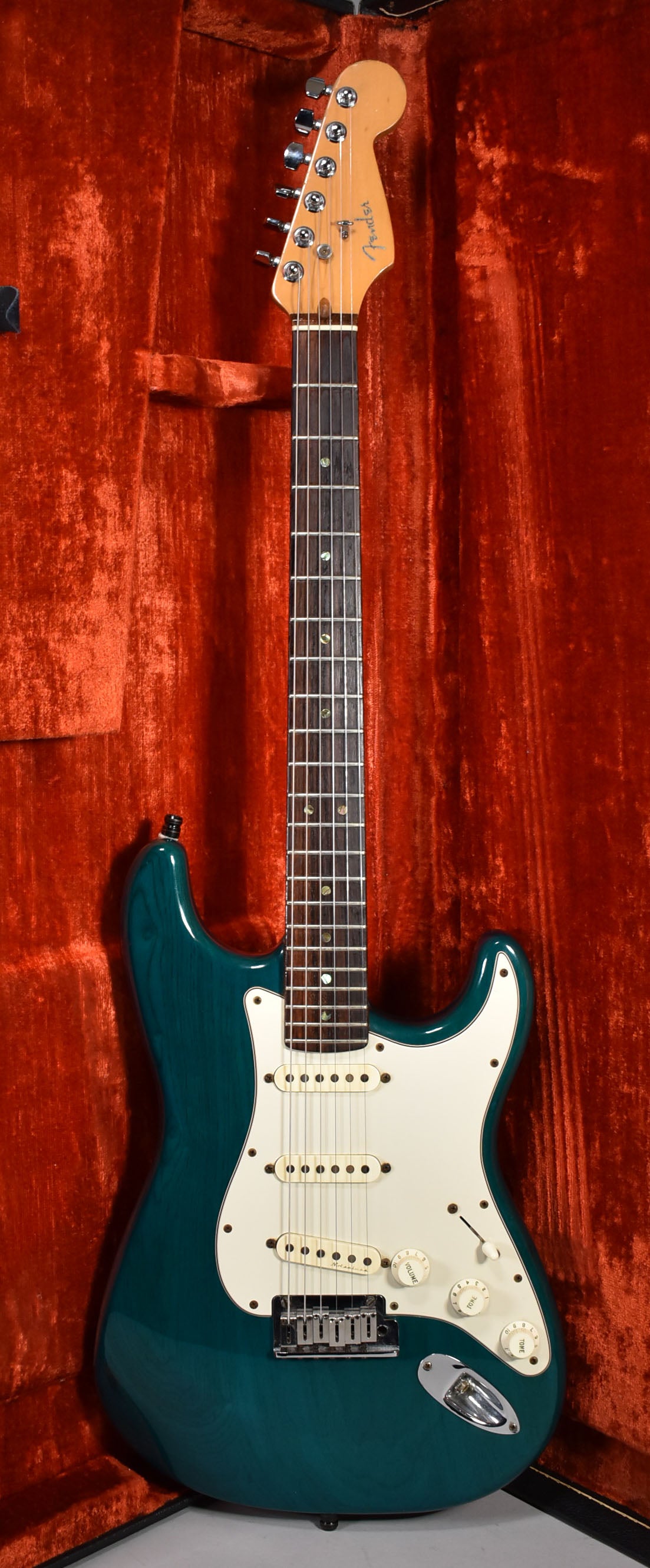 Full-length view of a transparent teal Stratocaster with a white pickguard, three cream pickups, and chrome hardware, resting in a red velvet case.