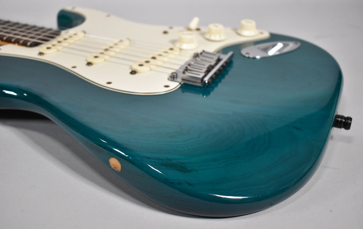 The lower bout of a transparent teal Stratocaster, showing wood grain, a chrome bridge, cream pickguard, and a small ding on the side.