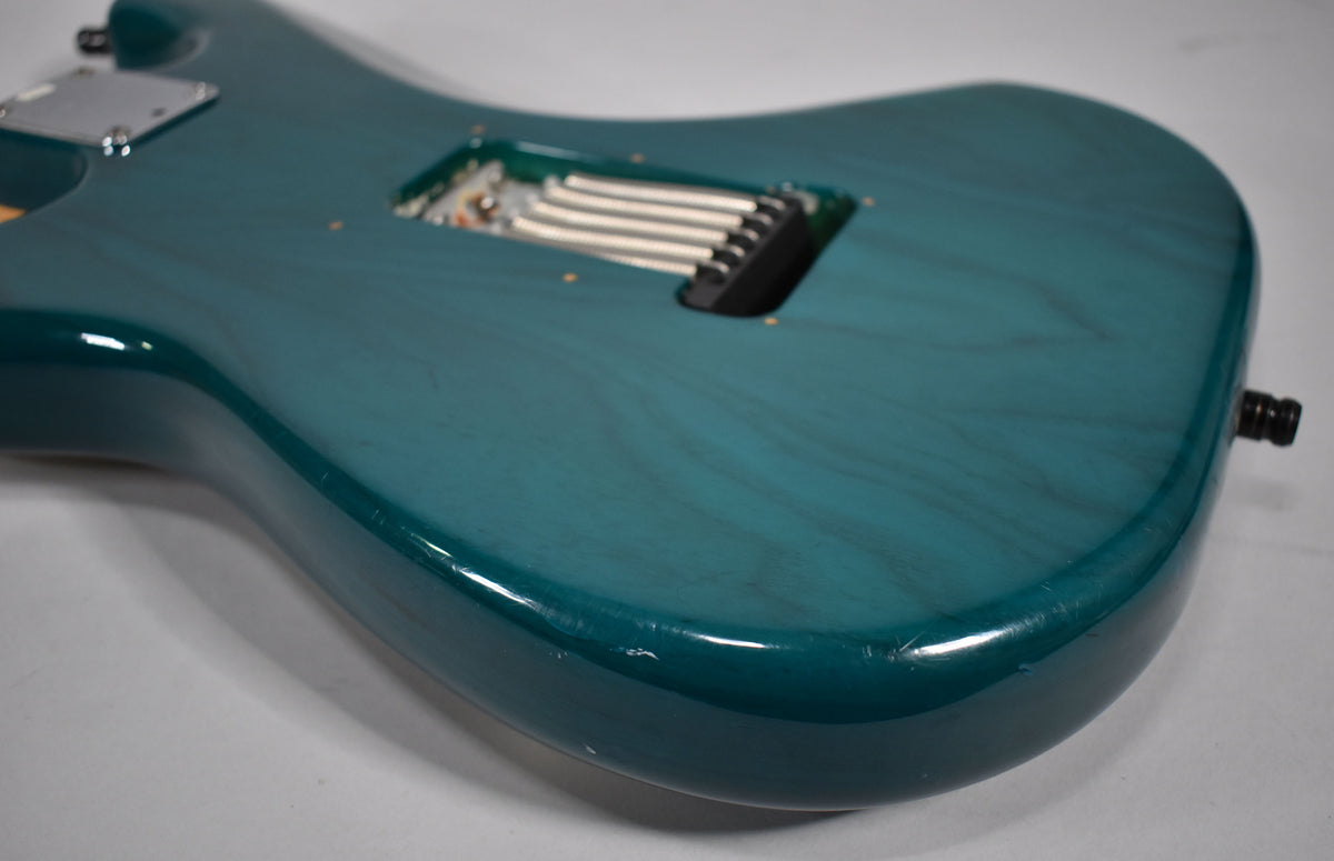 The back of a transparent teal guitar body, showing the tremolo spring cavity and some light scratches.