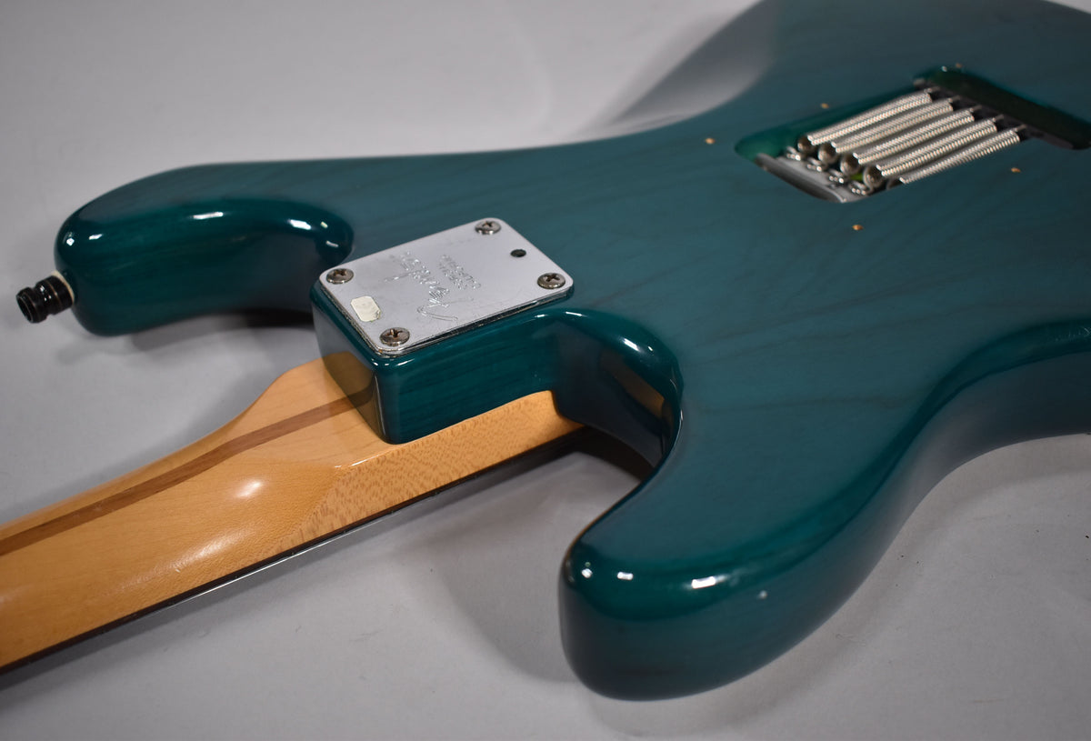 The guitar's transparent teal body, showing wood grain, has a chrome neck plate and a tremolo spring cavity on the back.