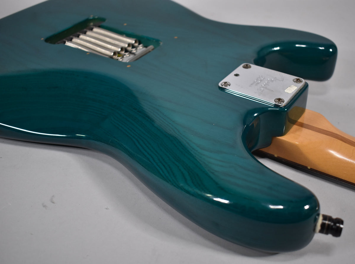 The back of the transparent teal Stratocaster shows the tremolo spring cavity, neck plate, and output jack, with visible wood grain.