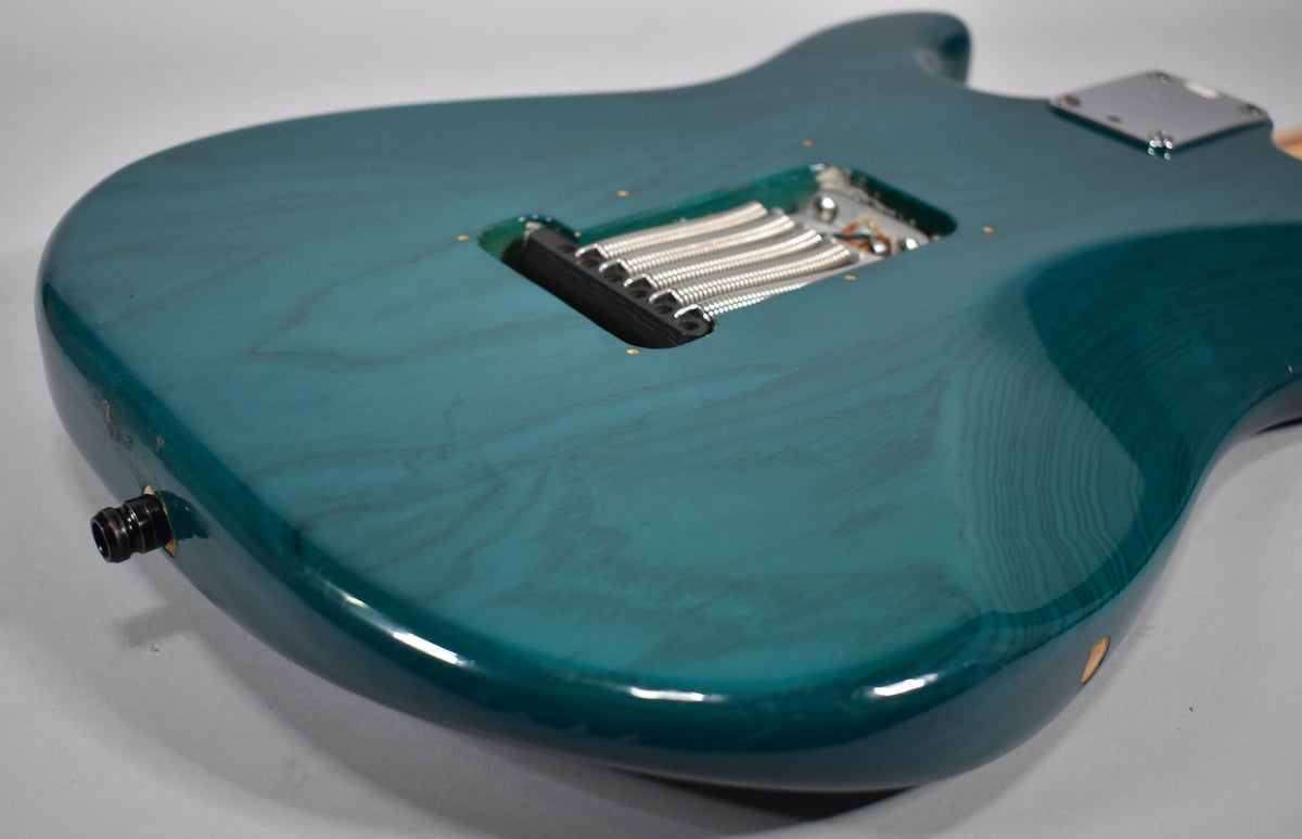 The guitar's transparent teal back shows wood grain, a tremolo spring cavity, and a black output jack with minor wear.