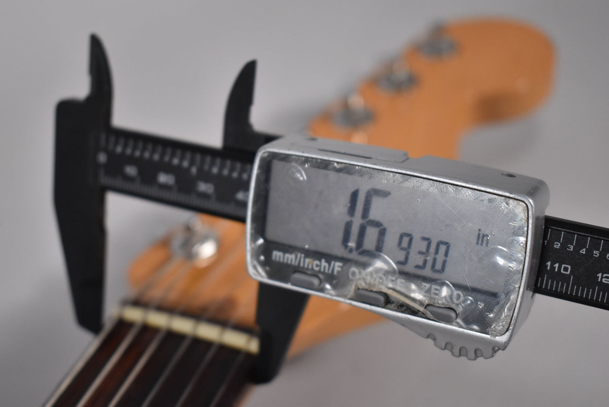 A digital caliper measures the guitar's nut width at 1.6930 inches, with the dark fretboard and light headstock blurred behind.