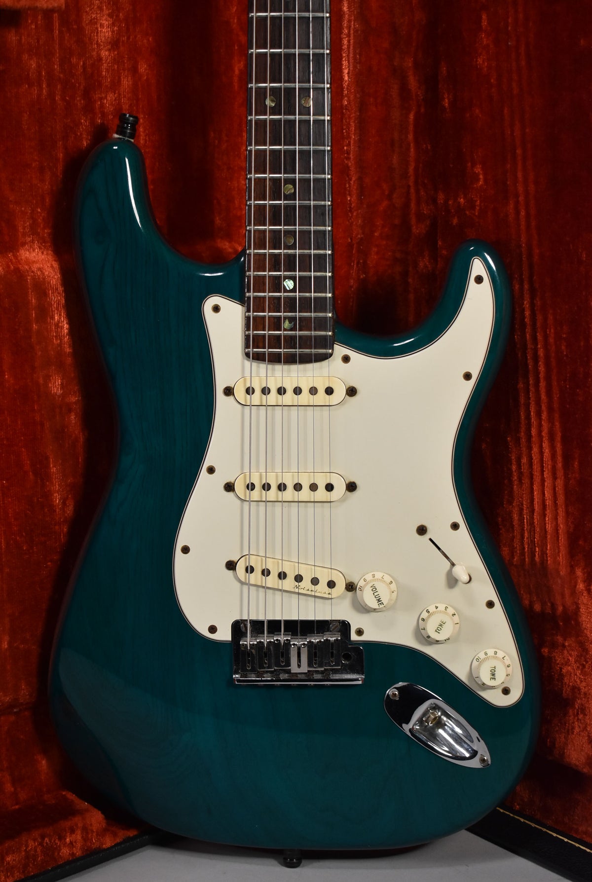 A transparent teal electric guitar, angled slightly right, shows its white pickguard, three cream pickups, and chrome hardware.