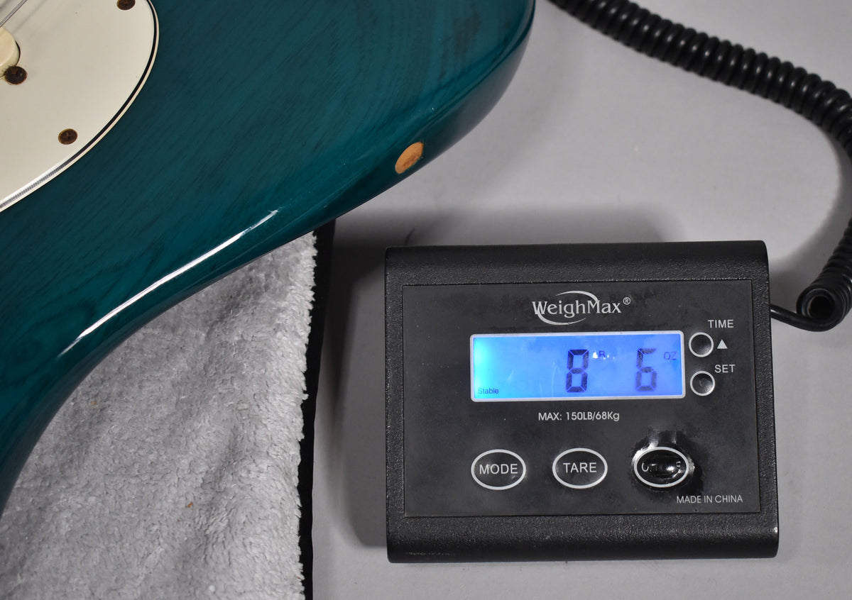 The guitar's lower bout in transparent teal is shown on a scale displaying 8 lbs 6 oz, with a visible scratch on the body.