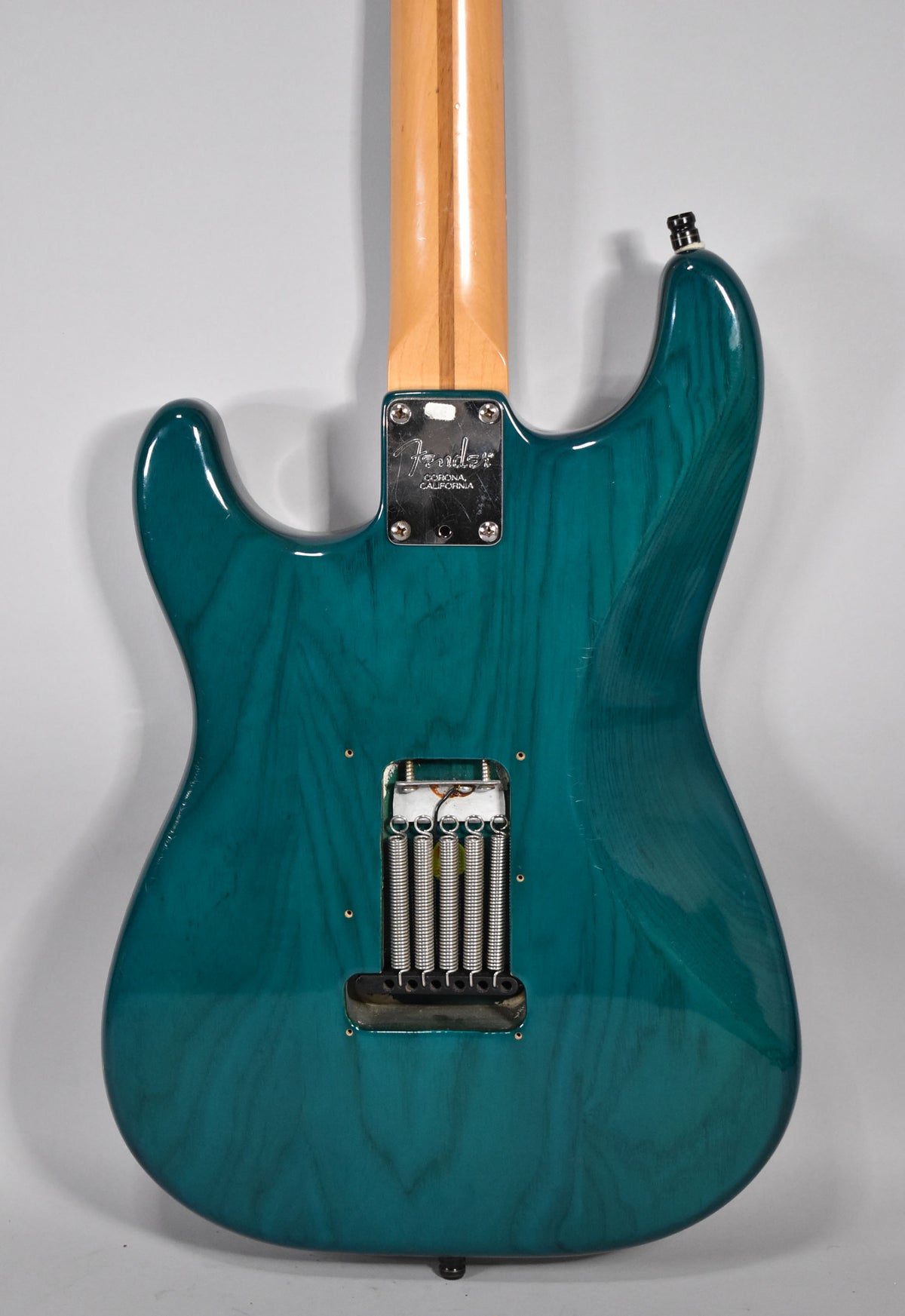 Rear view of a transparent teal Stratocaster, showing the wood grain, neck plate, and tremolo springs in the open cavity.