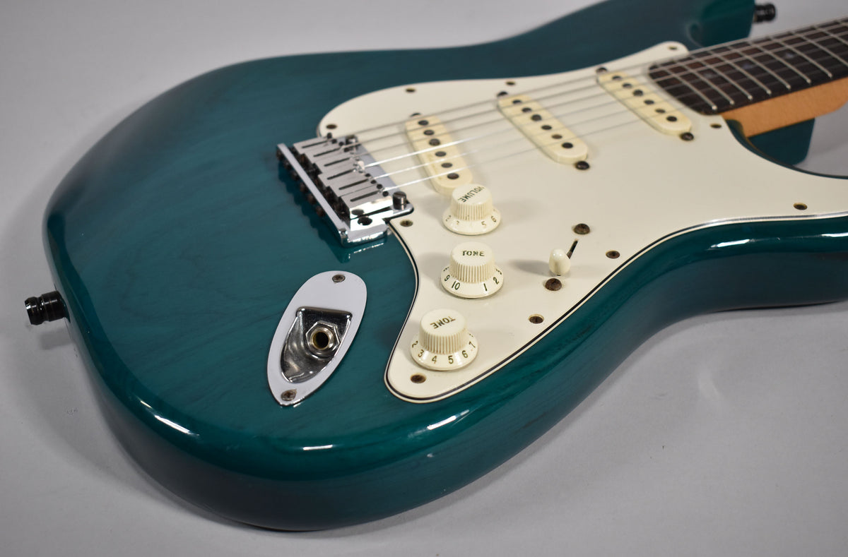 The transparent teal Stratocaster body, with visible wood grain, features a white pickguard, three cream single-coil pickups, and three cream control knobs.