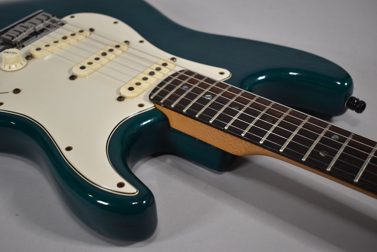 The teal guitar body and white pickguard are viewed from above, showing the bridge, three cream pickups, and fretboard.