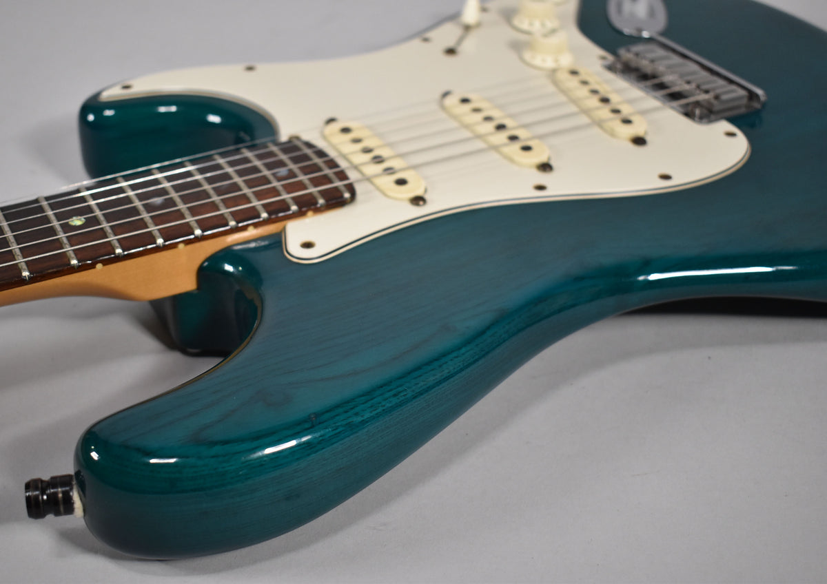 Close-up of the transparent teal guitar body, showing wood grain, white pickguard, three cream single-coil pickups, and chrome bridge.