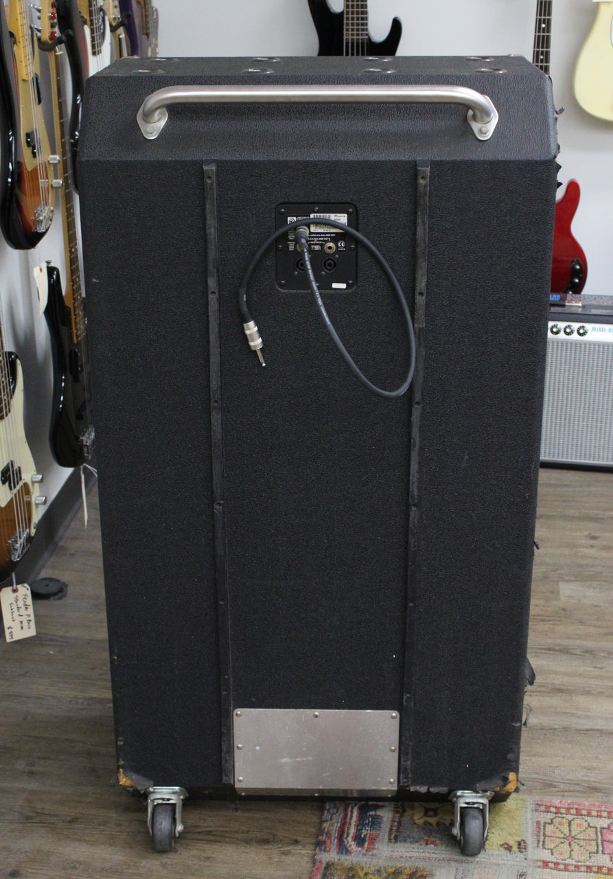 Rear view of the tall, black textured cabinet on casters, showing the input panel with a cable plugged in, and a metal kick plate.
