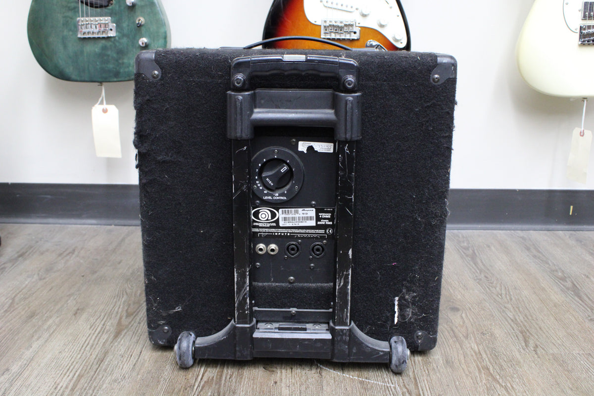 Rear view of a black carpet-covered speaker cabinet with a retractable handle, two wheels, and a control panel with a large knob and inputs.