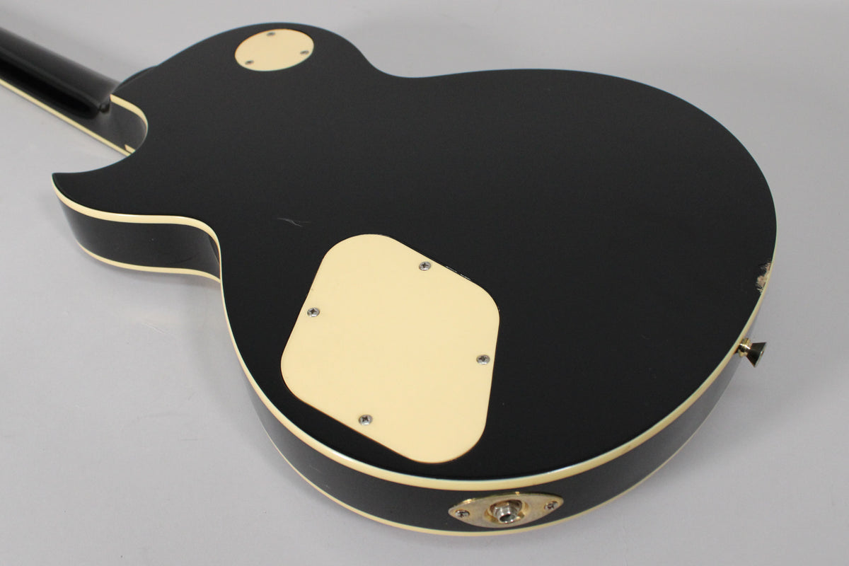 The back of a black gloss guitar with cream binding, showing control cavity covers and input jack, with some wear on the edge.