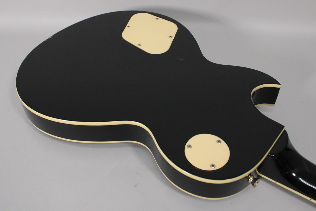 The back of a black gloss Les Paul-style guitar, showing cream binding, two cream control cavity covers, and some minor wear.