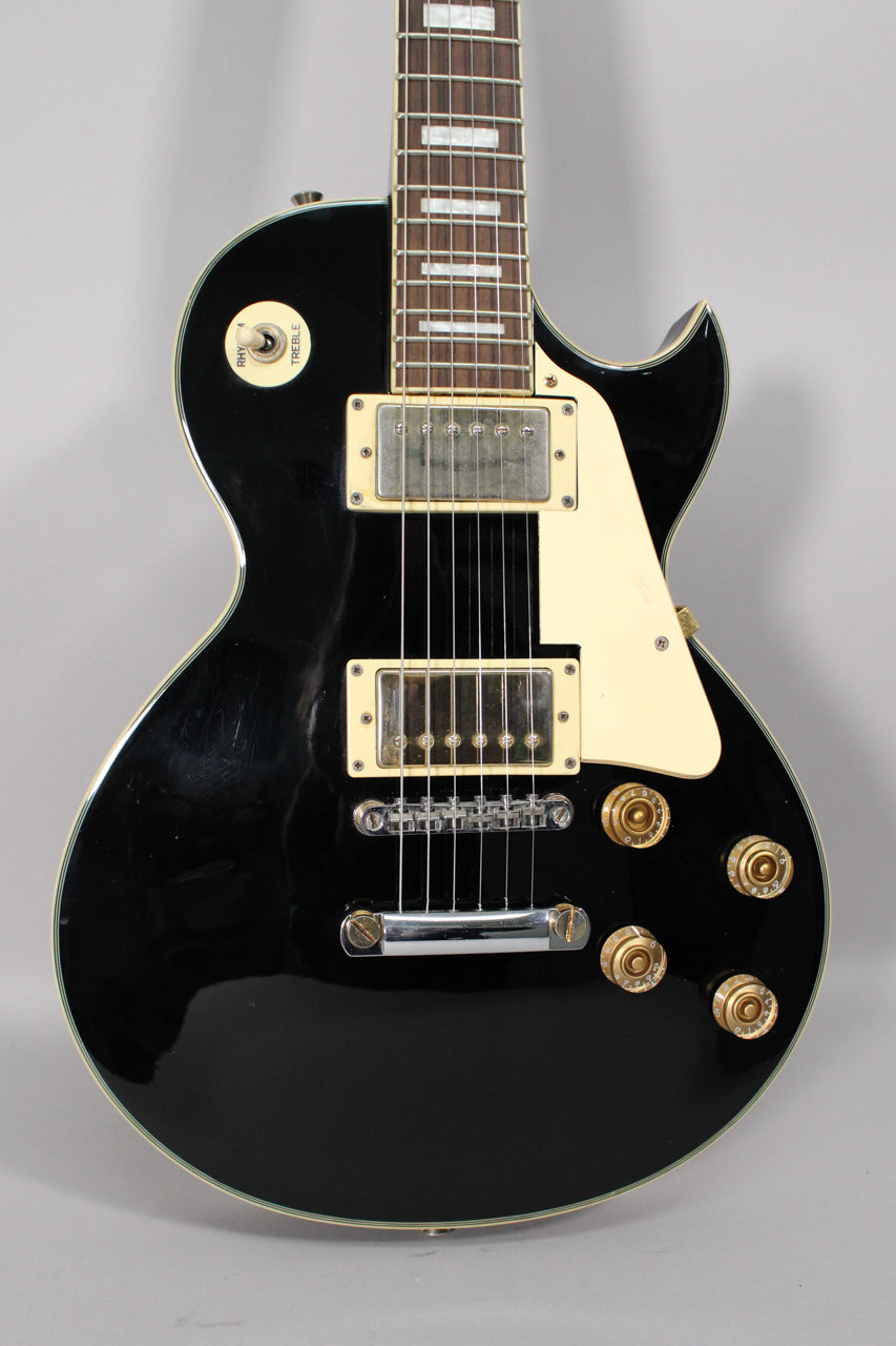 The black gloss guitar body is framed from the neck joint down, showing cream humbucker pickups, pickguard, and gold knobs.