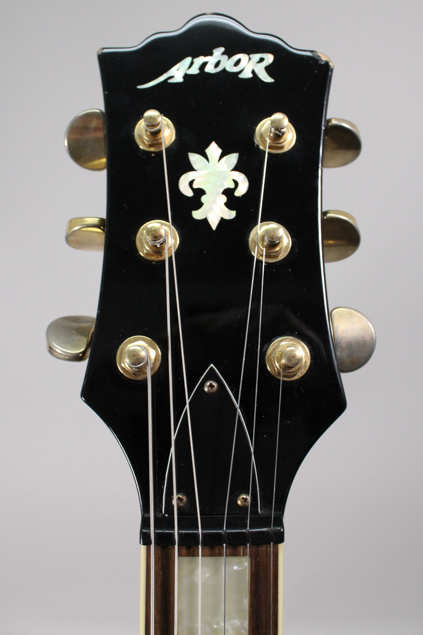 Close-up of the black gloss headstock with gold tuners, mother-of-pearl fleur-de-lis inlay, and minor finish wear on the top edge.