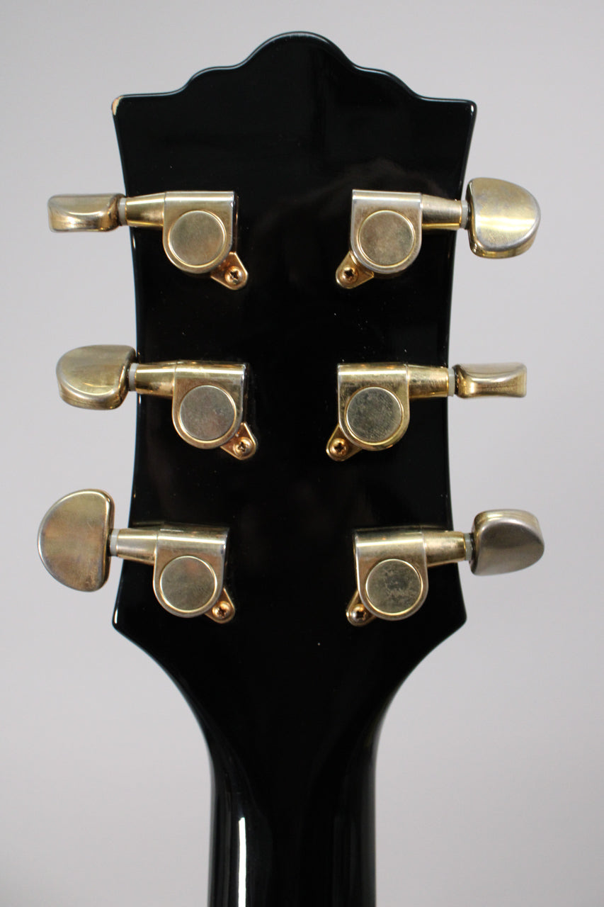 Rear view of the black gloss headstock with six gold-plated tuners, showing some wear and a chip on the top left edge.