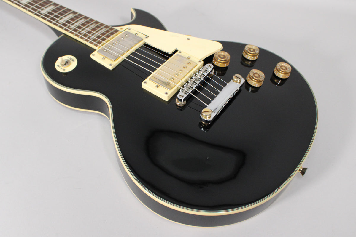 The black gloss Les Paul copy is angled from the neck, showing cream binding, pickups, pickguard, and four gold control knobs.