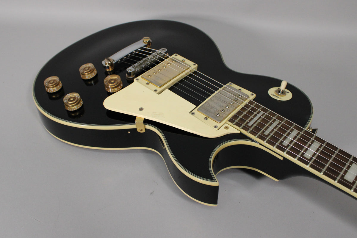 The black gloss Les Paul copy is angled from the upper body, showing cream binding, pickguard, and trapezoid fretboard inlays.