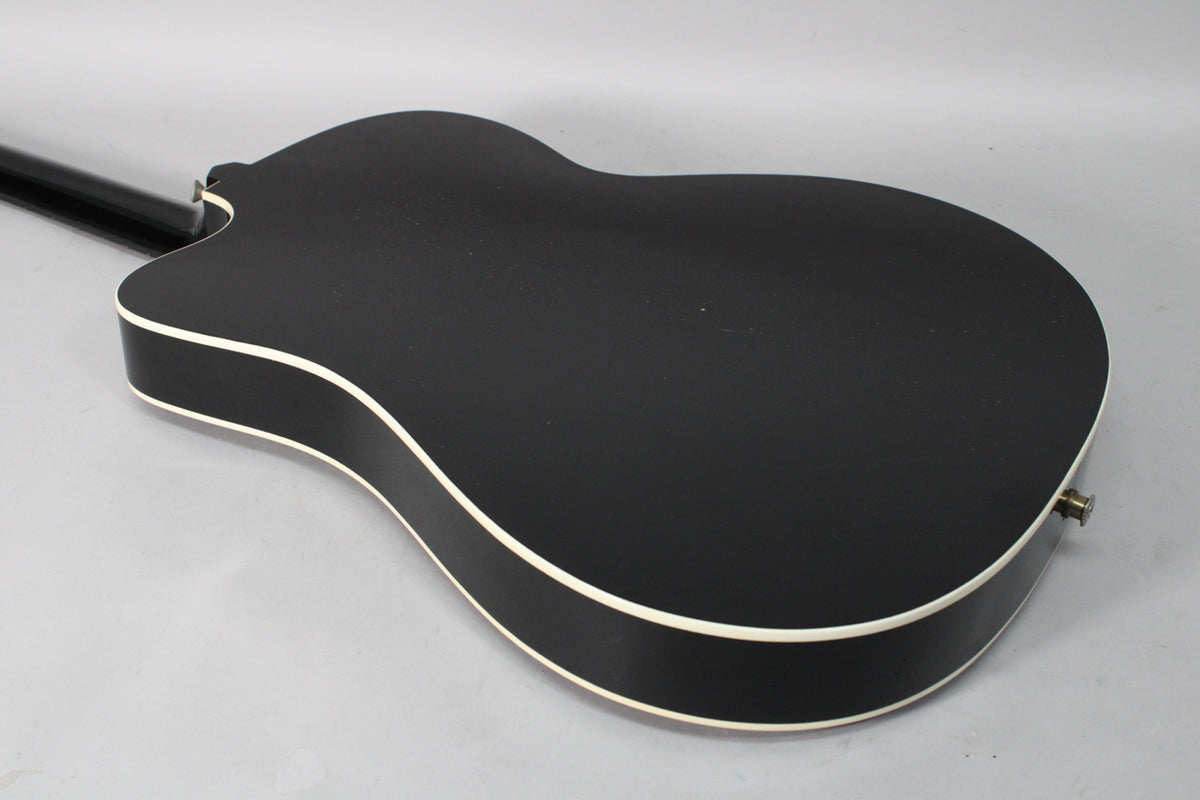The back of a black thinline acoustic-electric guitar, showing its matte finish, white binding, and a strap button/output jack.