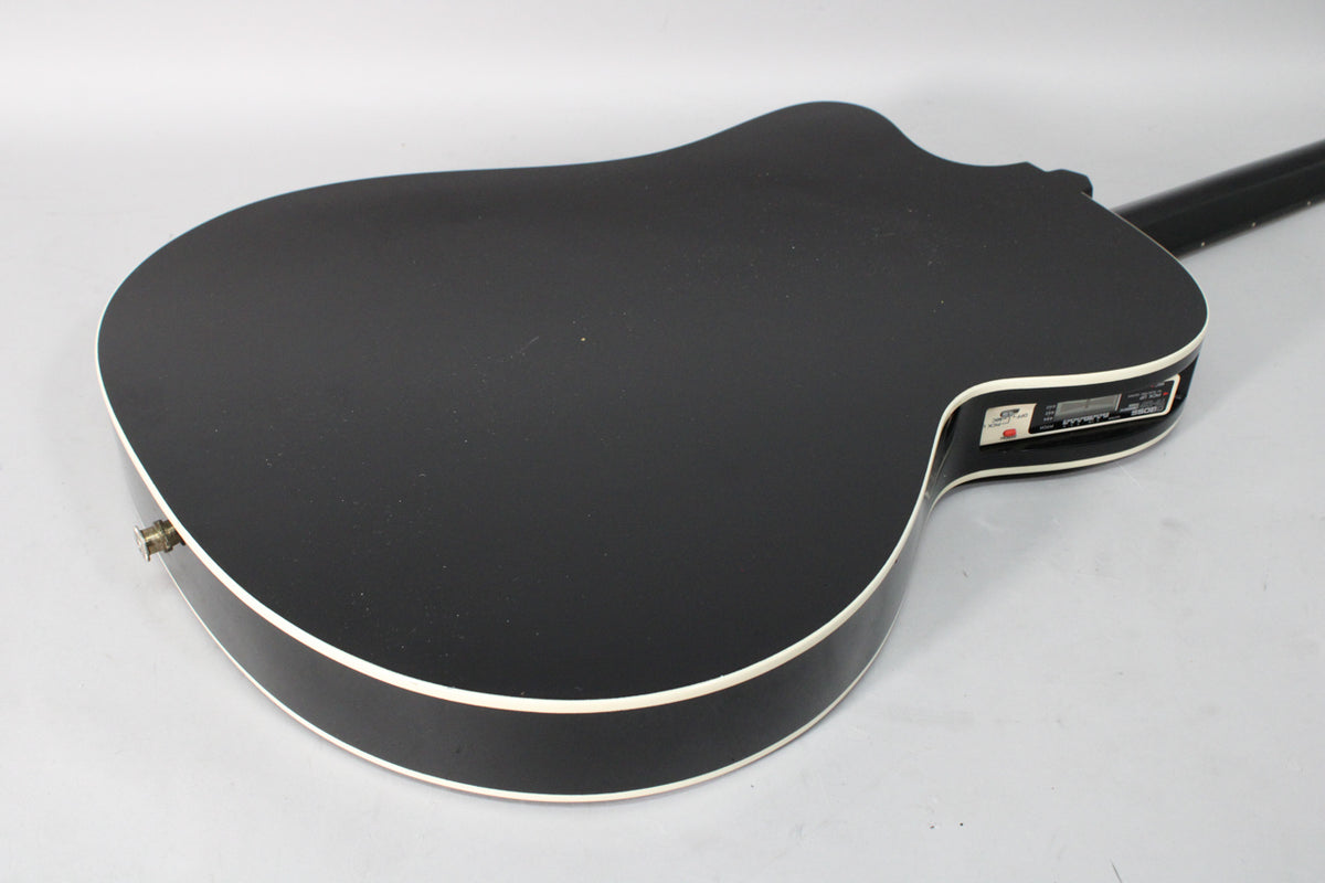 The black matte thinline acoustic-electric guitar's back and side are visible, with white binding and an active preamp on the upper bout.