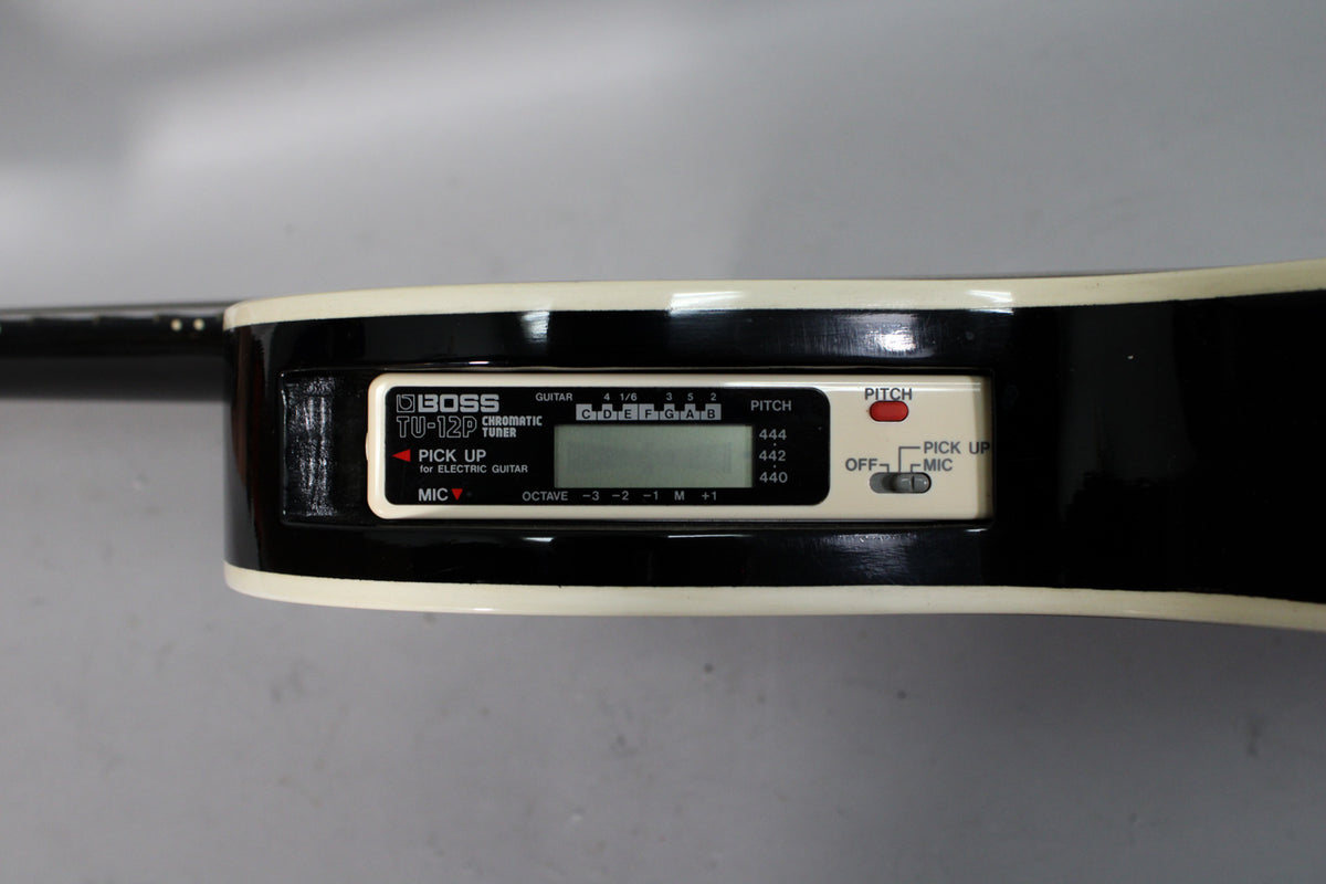 Close-up of the guitar's upper bout, showing a built-in tuner with a black LCD screen and white controls, set into the glossy black body.