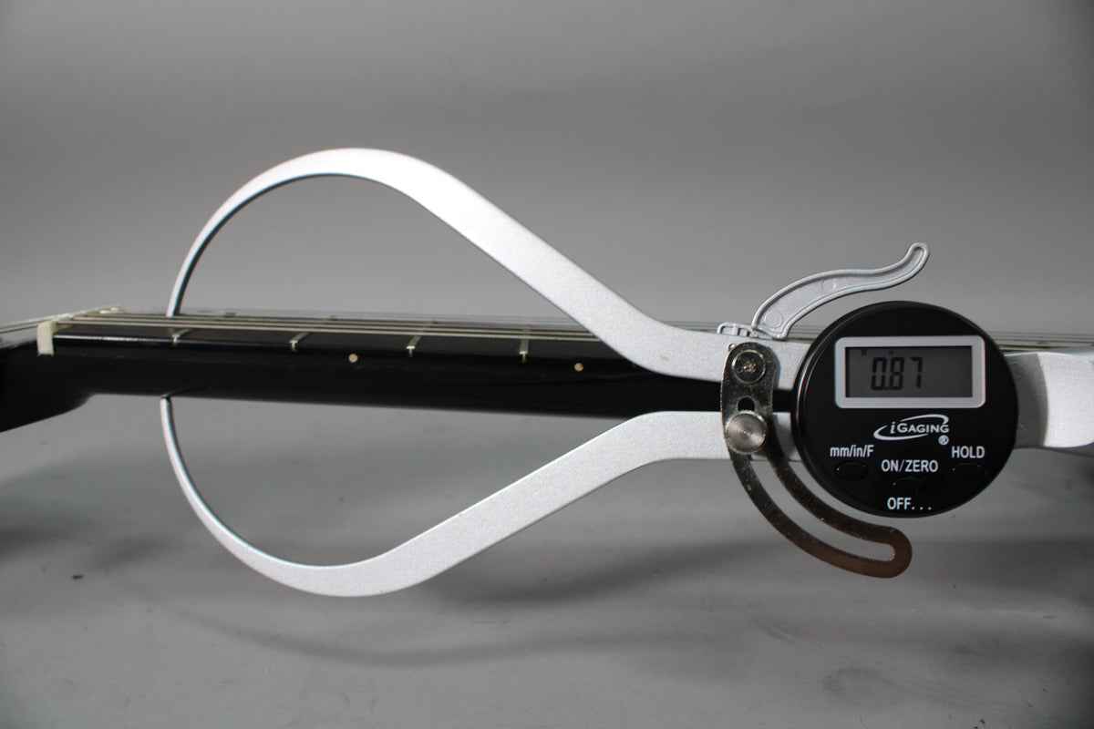 A digital caliper measures the string height at the 12th fret of the black guitar neck, displaying 0.87mm.
