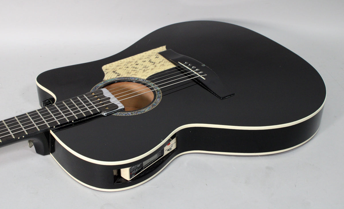 Black acoustic-electric guitar, angled from the neck, showing the cutaway, soundhole with abalone rosette, and side-mounted preamp.