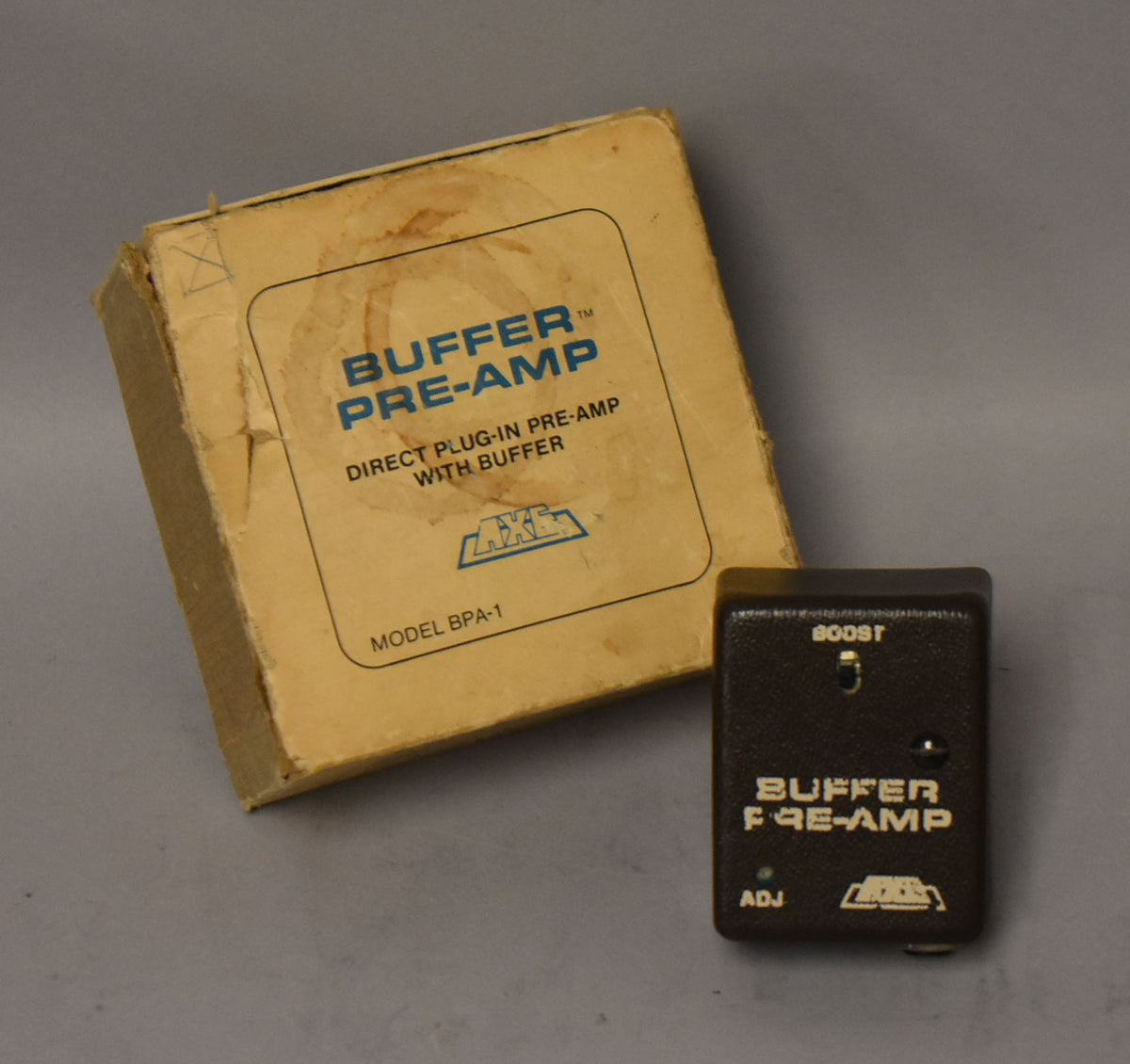 A dark brown, textured buffer pre-amp pedal with a single knob and switch, next to its worn, light brown cardboard box.