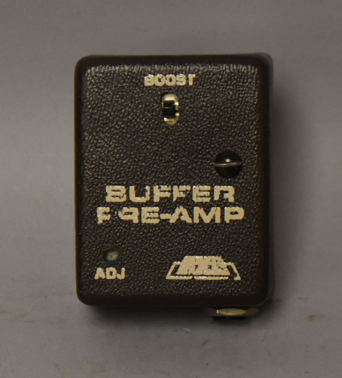 Top-down view of a dark brown, textured buffer pre-amp pedal with a small toggle switch, a screw, and a tiny green light.