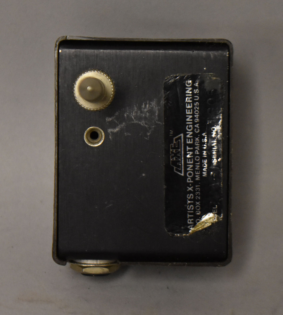 Top-down view of a rectangular black metal pedal with a cream knob, a small gold jack, and a worn black label.