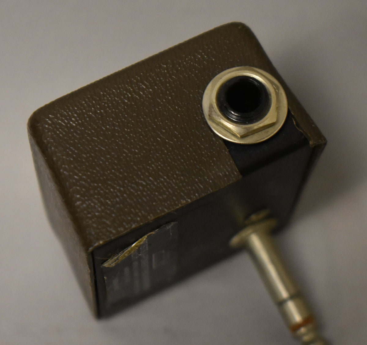 Top-down view of the olive-green textured pedal, showing a brass input jack and a connected cable, with some peeling on the side.