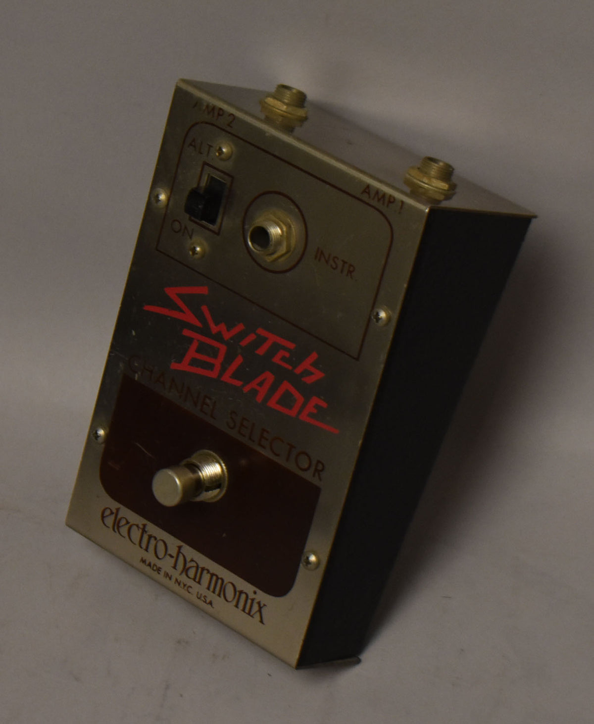 Gold-finished metal pedal with black sides, showing a toggle switch, three jacks, and a footswitch, with some surface wear.