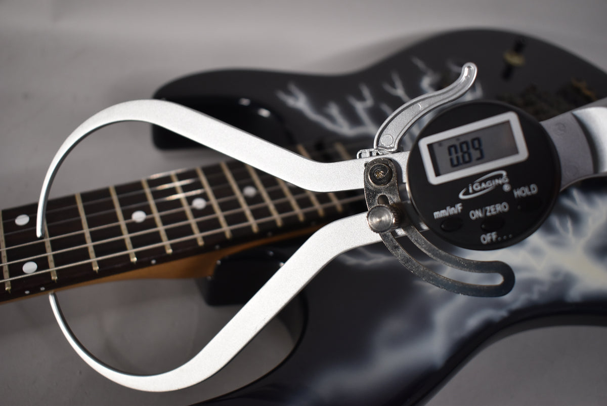 A digital caliper measures the fret height at 0.89mm on the dark fretboard of the black guitar with white lightning graphics.
