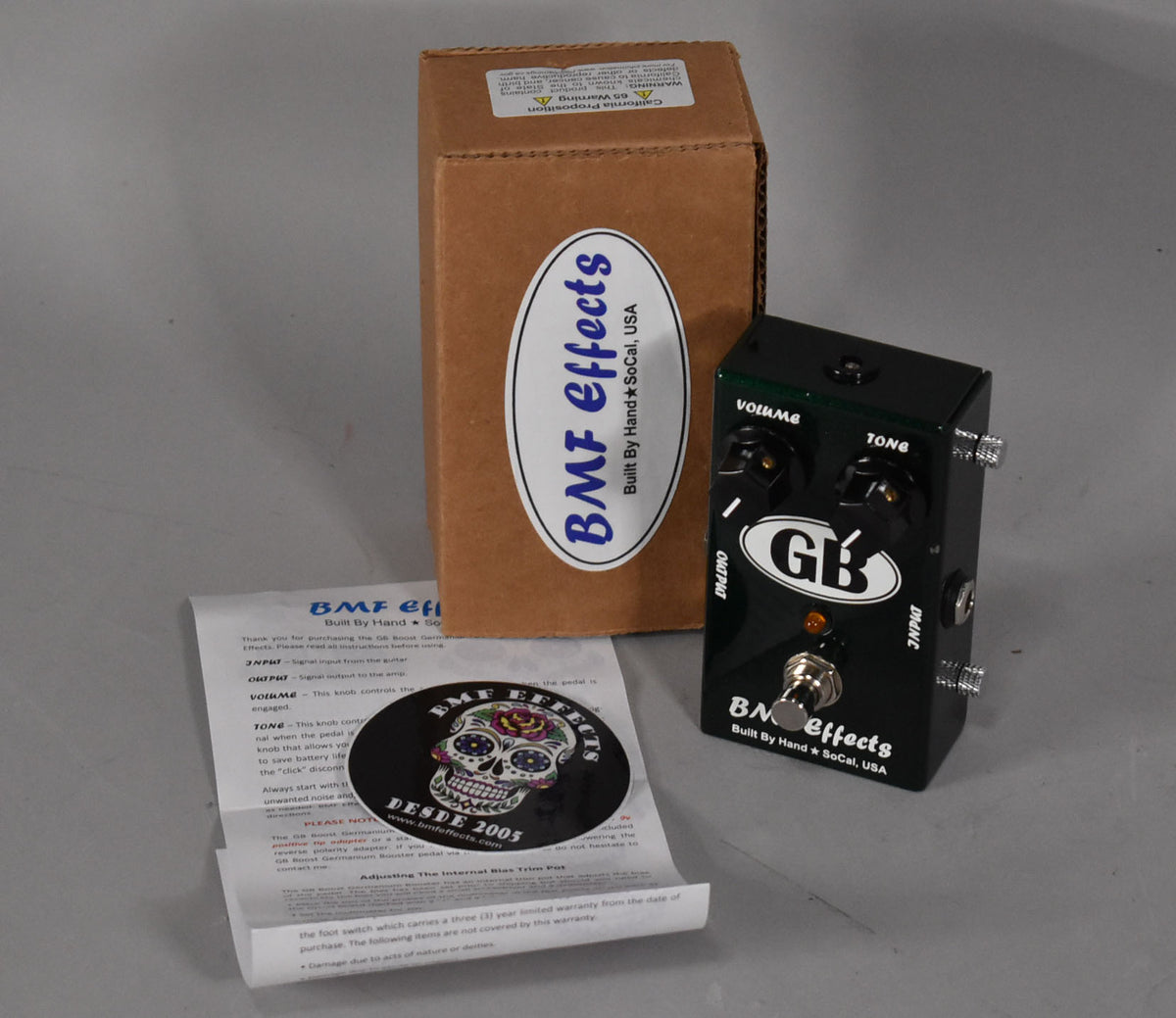 A dark green boost pedal with two black knobs, a footswitch, and an orange indicator light, next to its box and manual.