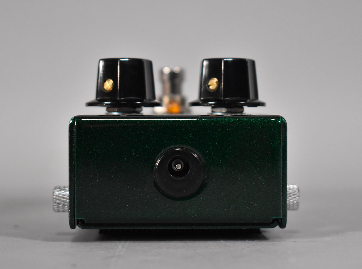 The dark green metallic pedal is viewed from the front, showing the power input and two black control knobs with gold indicators.