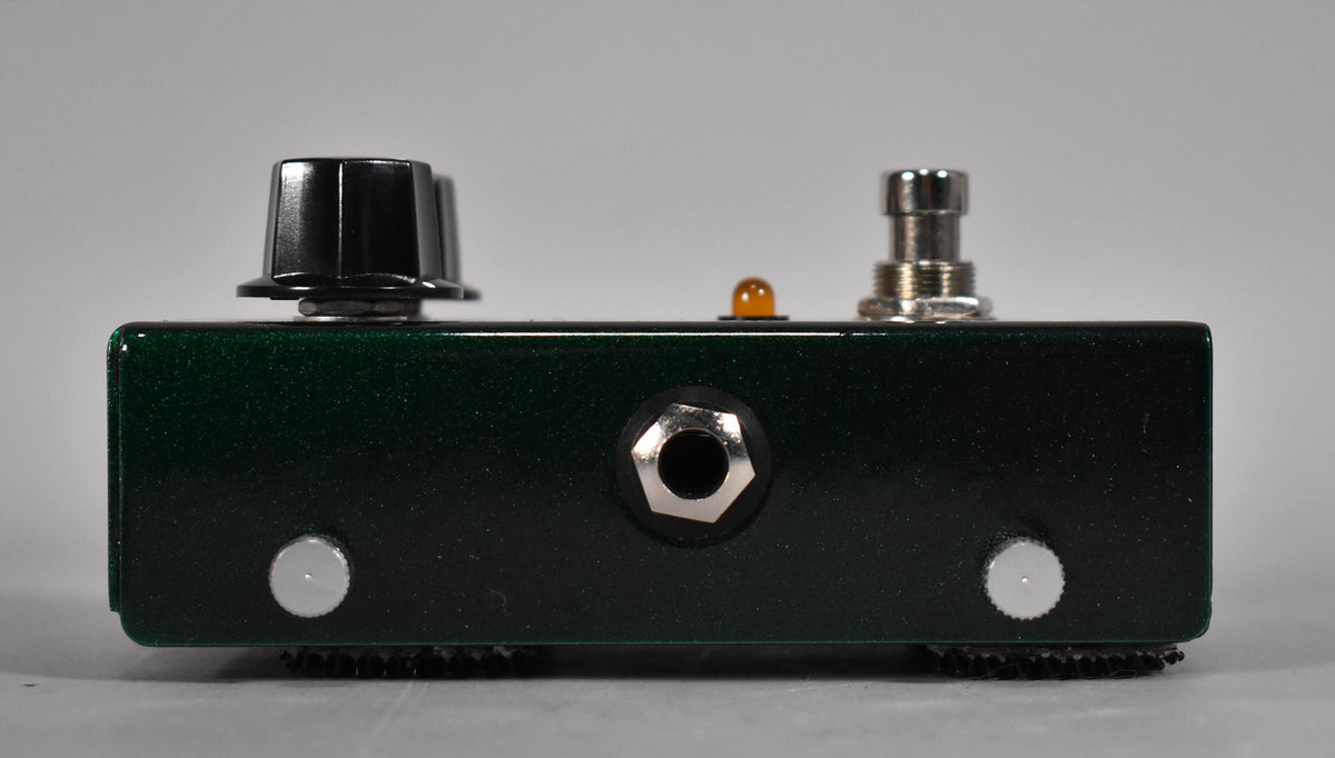 The dark green metallic pedal is viewed from the side, showing a black knob, amber LED, footswitch, and input jack.