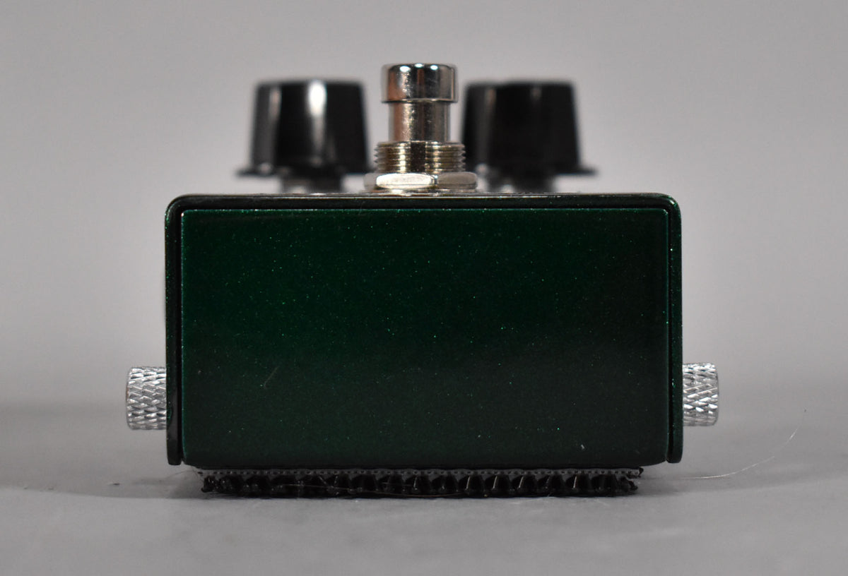 The dark green metallic pedal is viewed from the front, showing two knurled silver jacks and a silver footswitch.