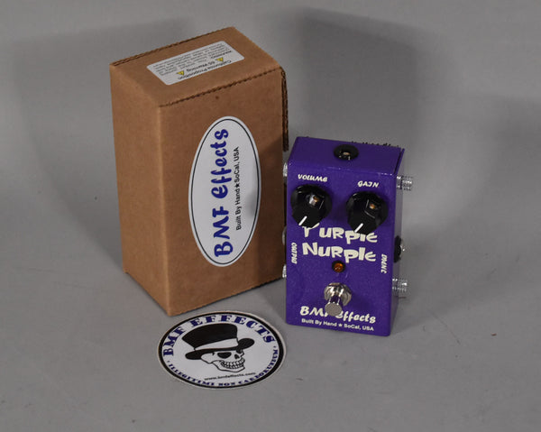 Purple guitar pedal with two black knobs, a red indicator light, and a footswitch, next to its brown cardboard box and a sticker.