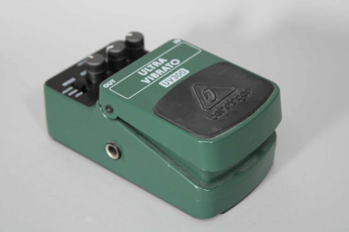 A dark green vibrato pedal, angled from the right, with a black rubber footpad and three black control knobs visible on the left.