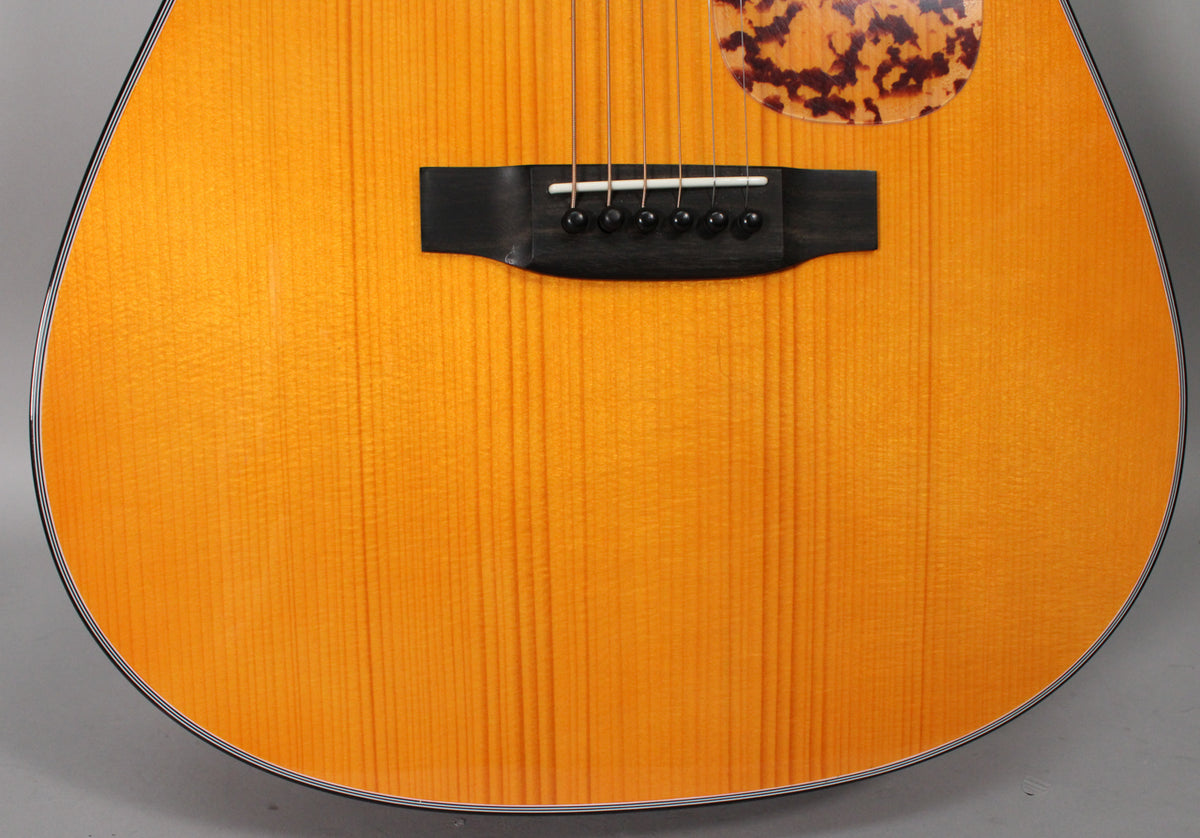 Close-up of the guitar's lower body, showing the light orange wood grain, black bridge, white saddle, black bridge pins, and tortoiseshell pickguard.