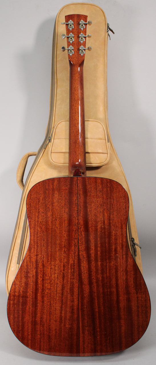 The guitar's dark reddish-brown mahogany back and neck are visible, with six chrome tuners on the headstock, resting in a tan gig bag.