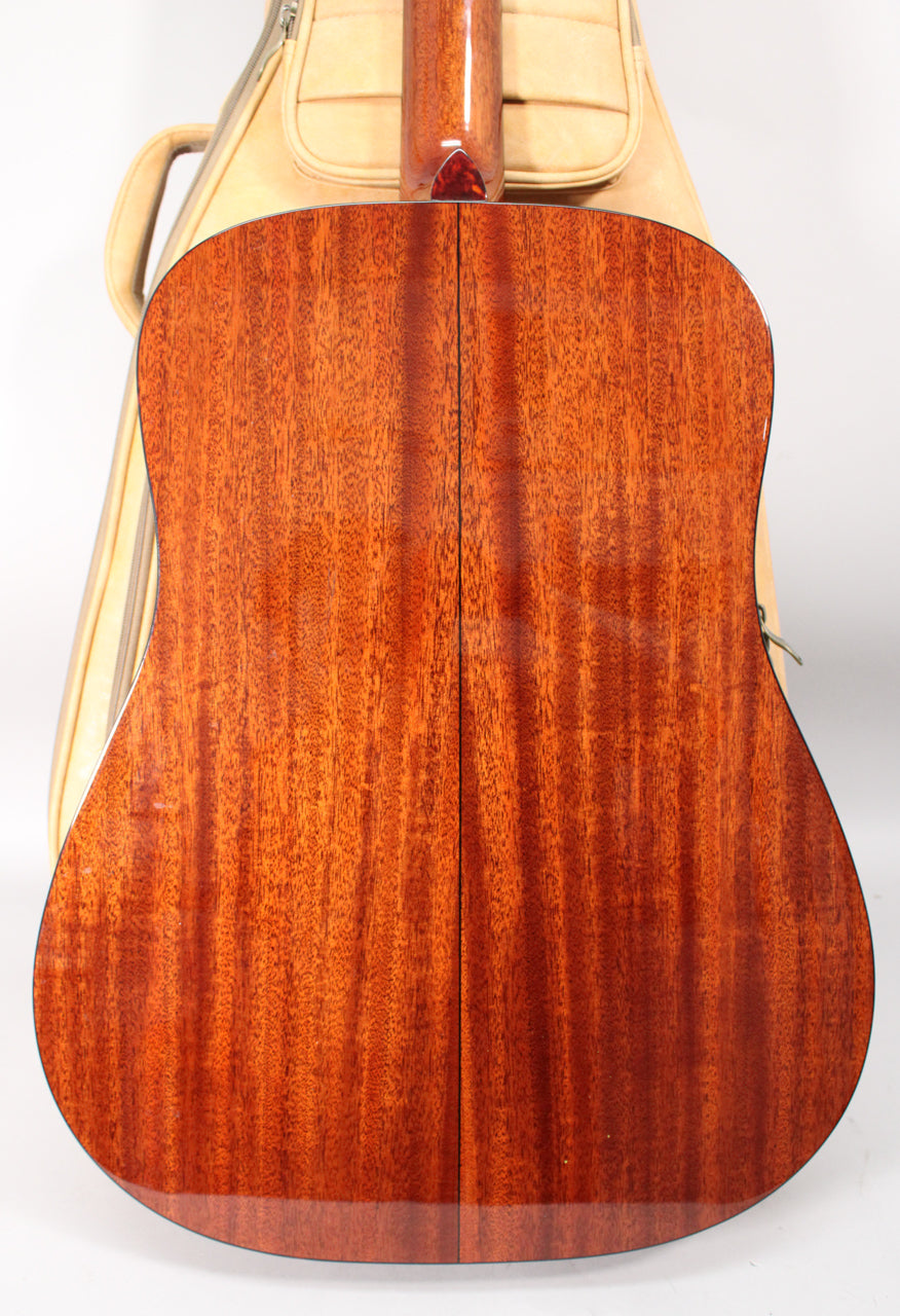 The guitar's glossy mahogany back, with visible wood grain and a subtle scratch, is centered against a beige gig bag.