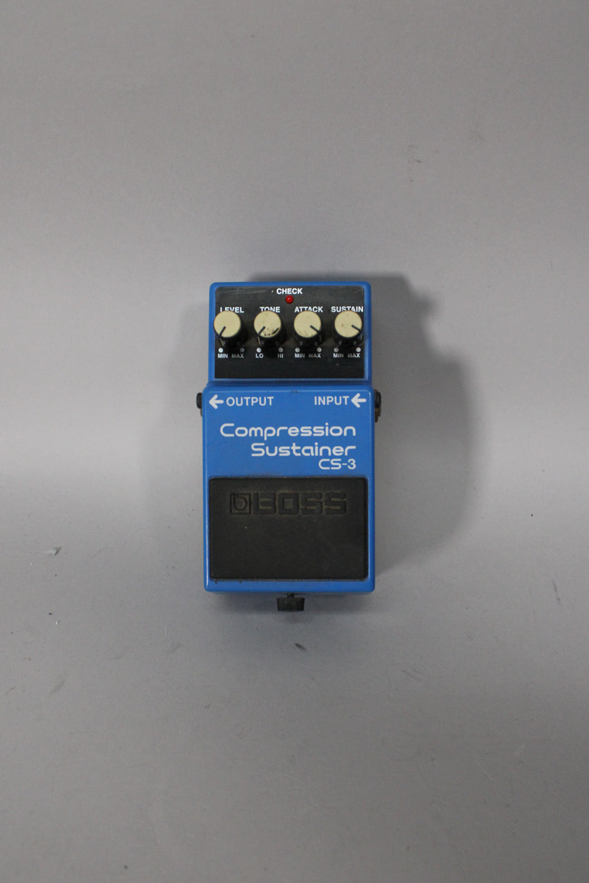 A top-down view of a blue Boss CS-3 pedal with four white control knobs and a red LED, showing some scuffs and dust.