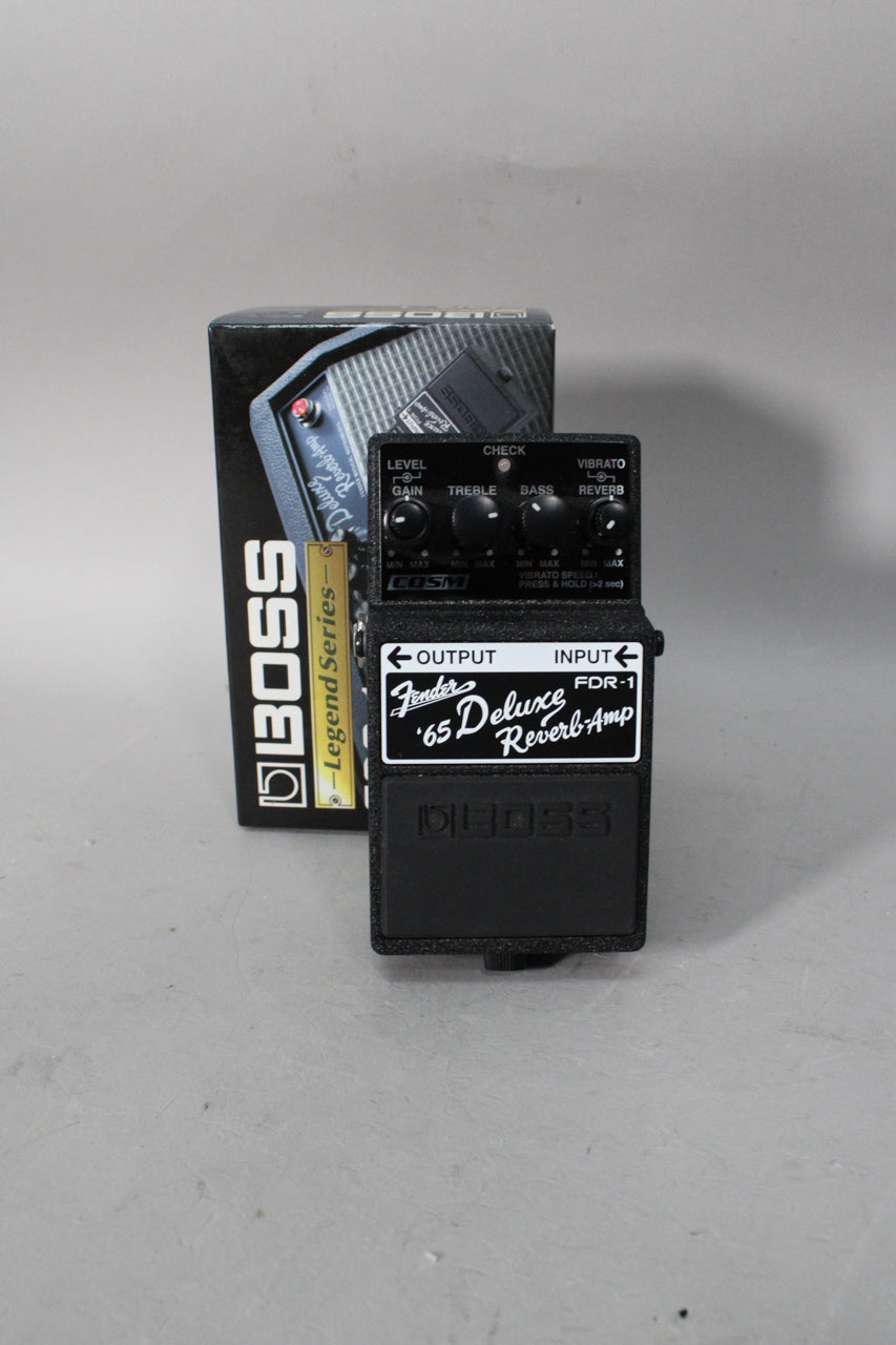 A black guitar pedal with a textured finish, viewed from above, showing its control knobs and footswitch, with its box behind it.
