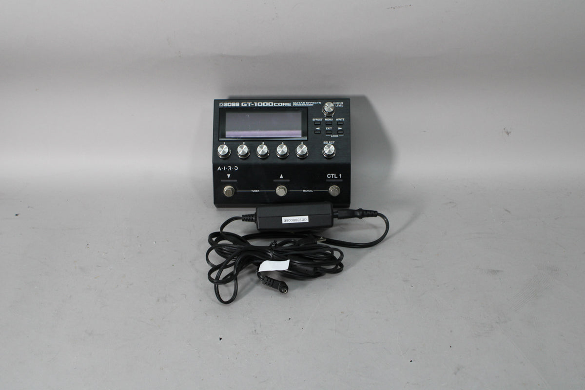 A black multi-effect processor with a large screen, six silver knobs, and three footswitches, shown with its power adapter.