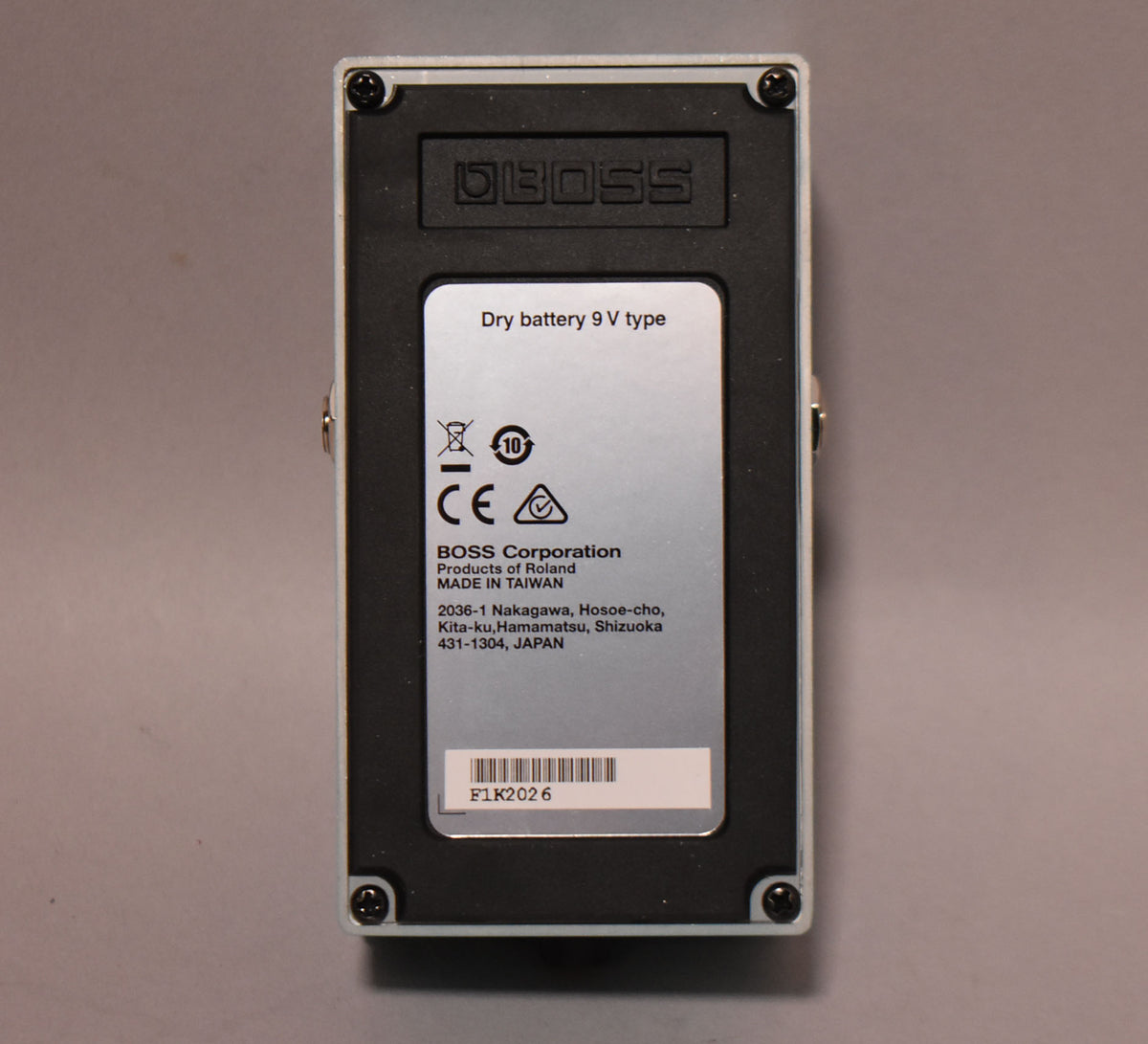 The pedal's silver-grey underside is shown, with a black battery compartment cover and a silver label with text.
