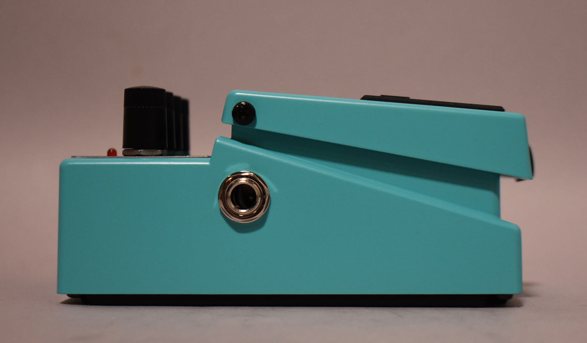 The turquoise pedal is viewed from the right side, showing a silver input jack, three black knobs, and a red indicator light.