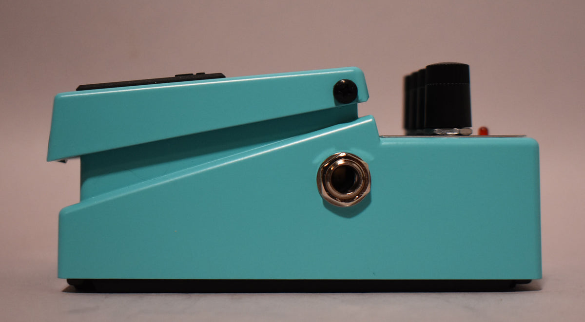 The turquoise pedal is viewed from the right side, showing a chrome input jack, three black control knobs, and a red LED.