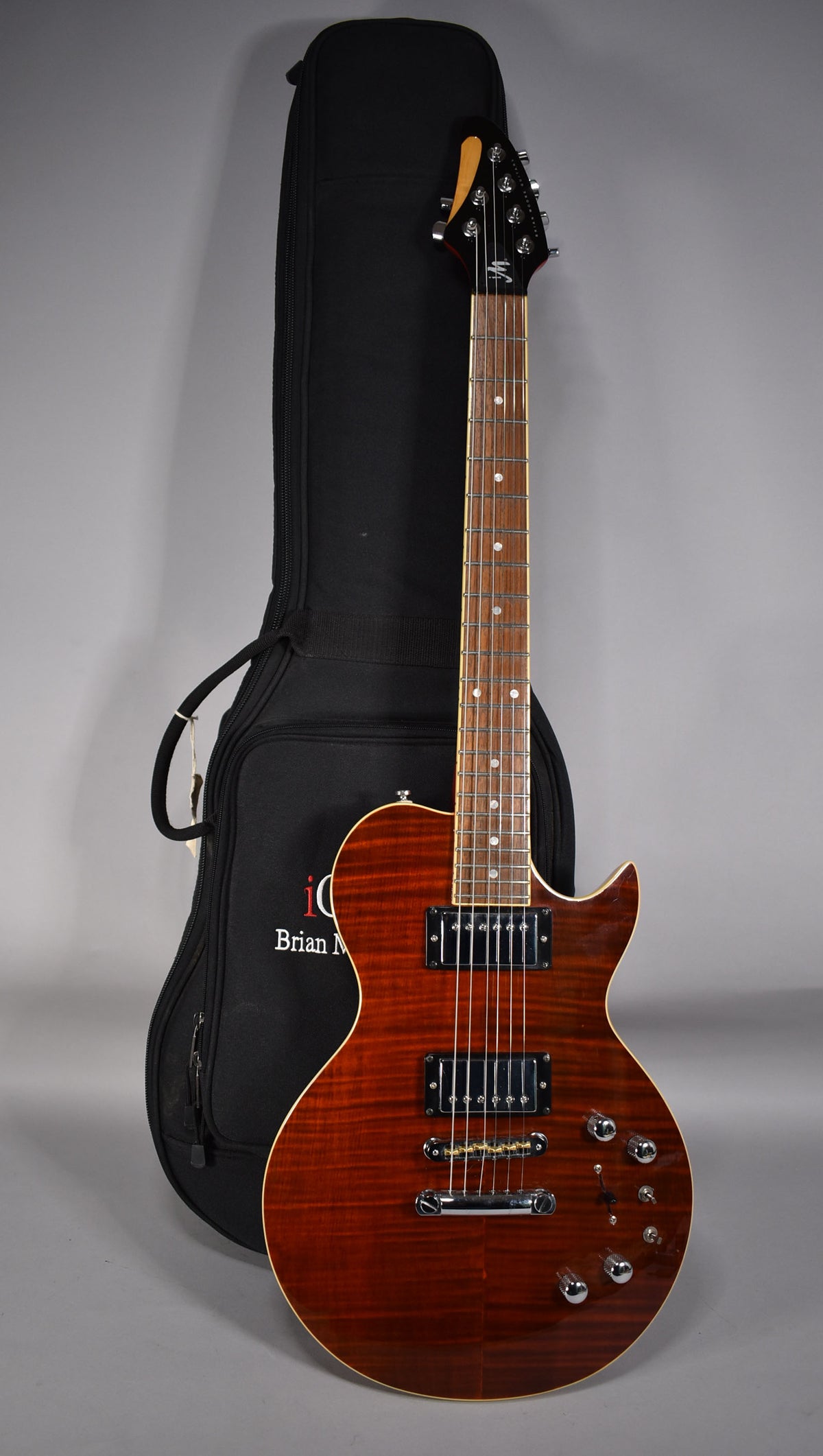 A full-length view of a glossy, cinnamon-burst electric guitar with chrome hardware and four control knobs, leaning against a black gig bag.